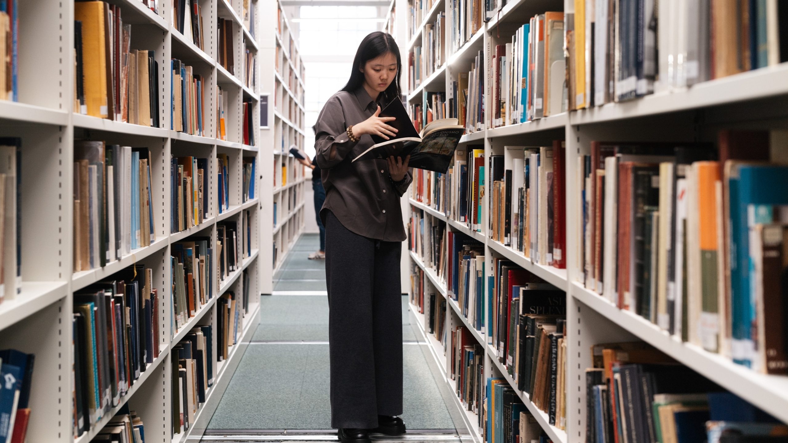 a female young student standing in a library between two rows of stacked books