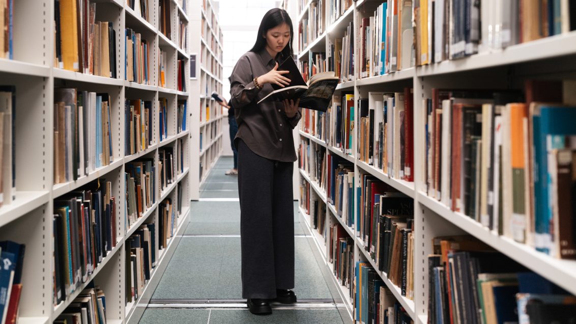a female young student standing in a library between two rows of stacked books