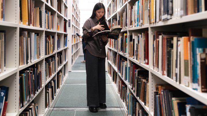 a female young student standing in a library between two rows of stacked books