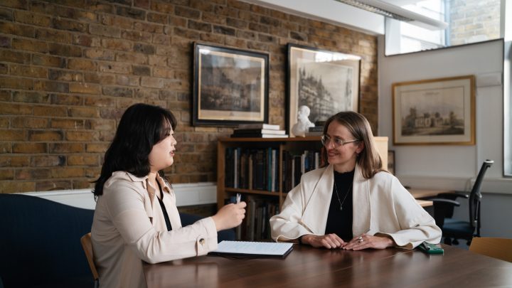 two women sitting at a table conversing with each others. There are some paitinings in frames nung on the walls behind them.