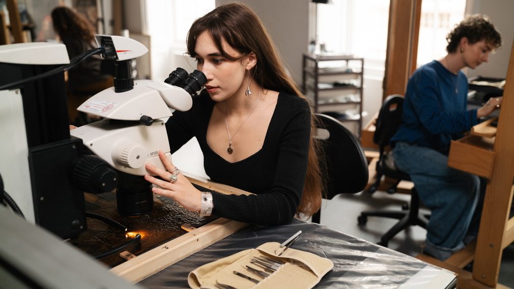 A woman looks through a microscope studying the surface of a painting. Behind her in the background, someone else works on an easel.