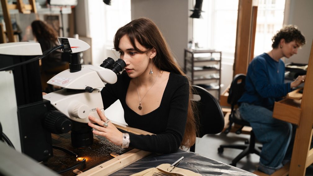 A woman looks through a microscope studying the surface of a painting. Behind her in the background, someone else works on an easel.