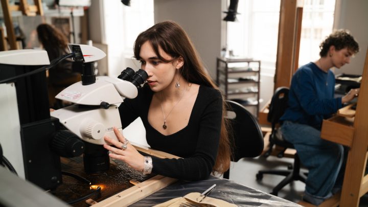 A woman looks through a microscope studying the surface of a painting. Behind her in the background, someone else works on an easel.