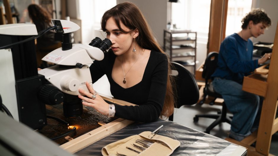A woman looks through a microscope studying the surface of a painting. Behind her in the background, someone else works on an easel.