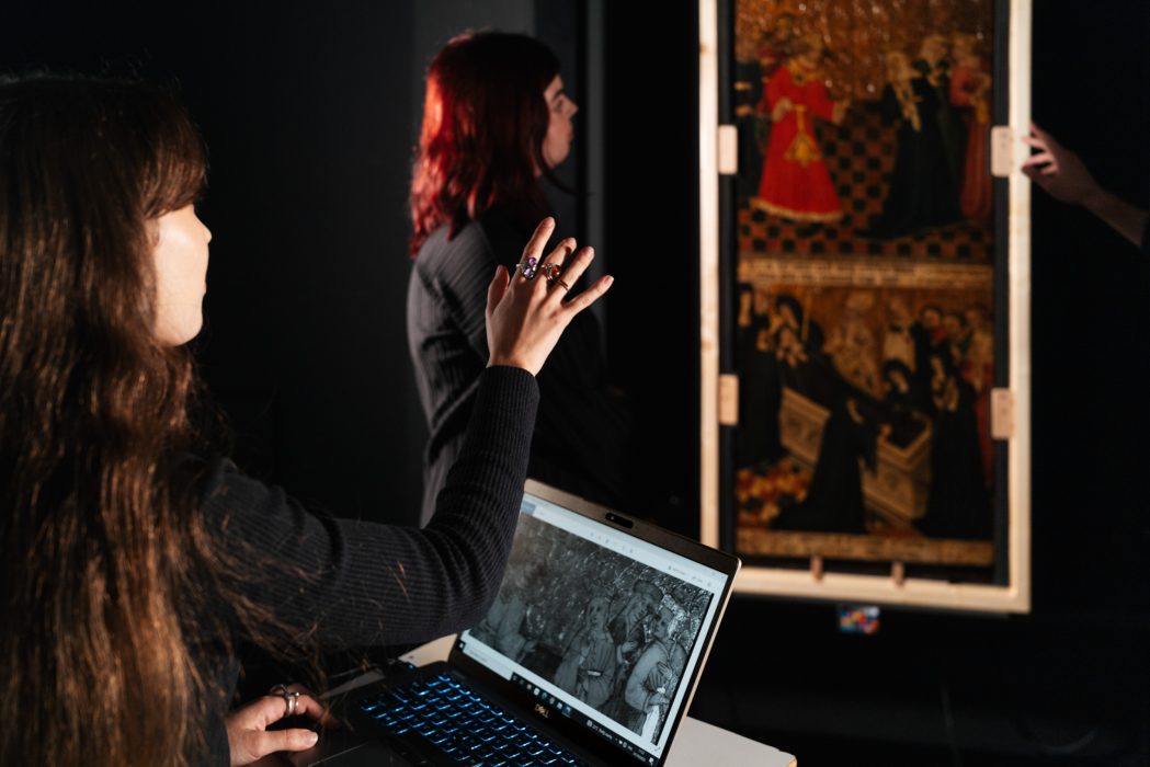In a dark studio, two women look at an artwork on the wall. One has a laptop open in front of her with a black and white photograph of the same artwork.