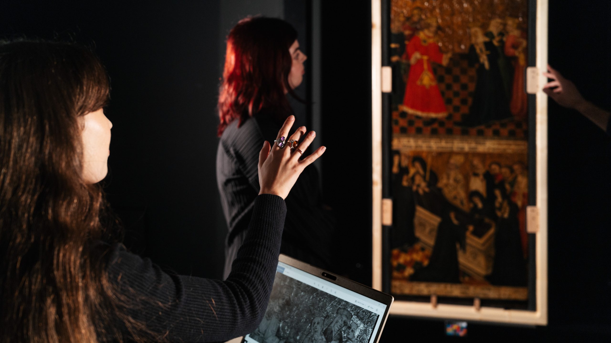 In a dark studio, two women look at an artwork on the wall. One has a laptop open in front of her with a black and white photograph of the same artwork.