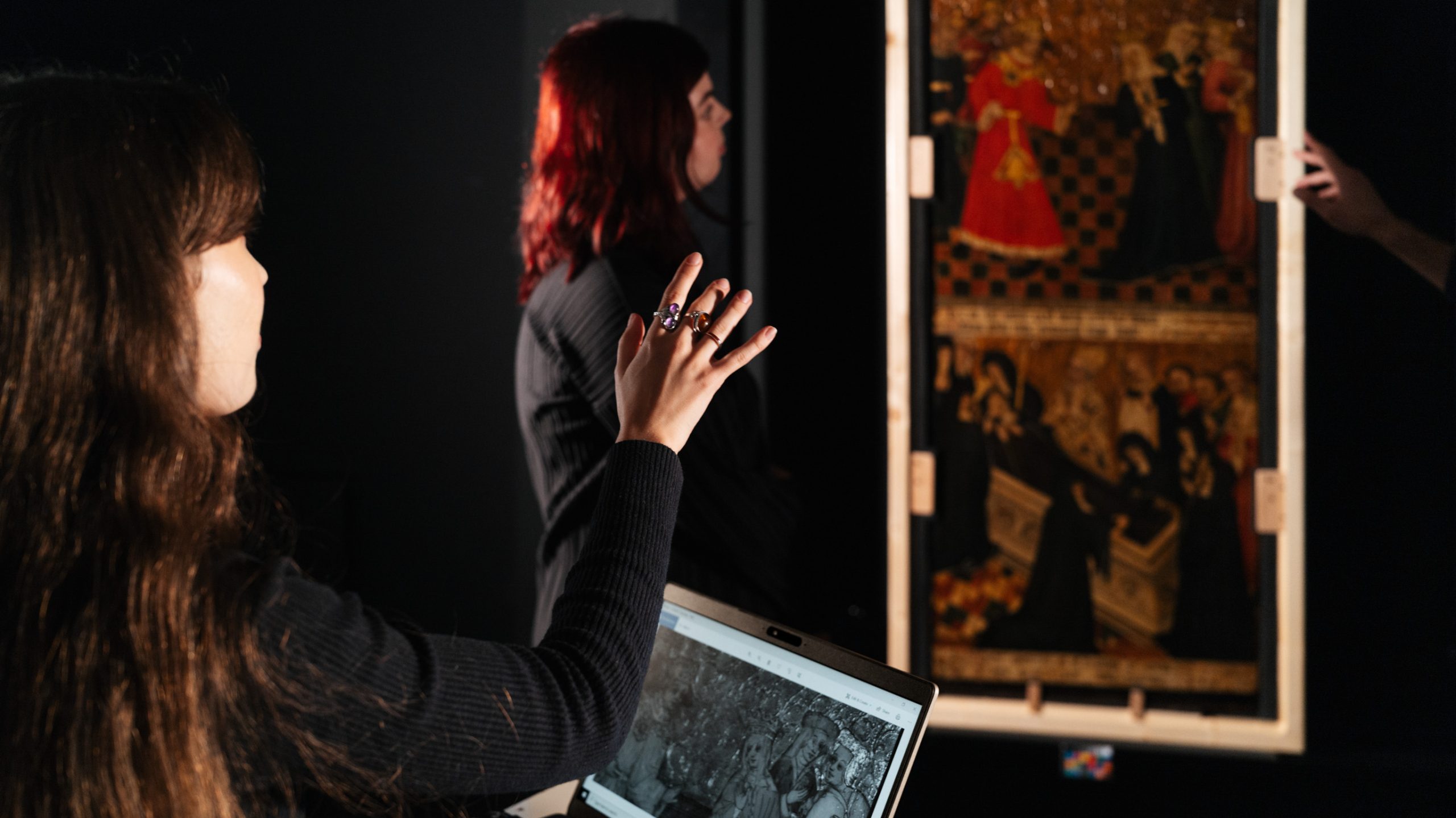 In a dark studio, two women look at an artwork on the wall. One has a laptop open in front of her with a black and white photograph of the same artwork.