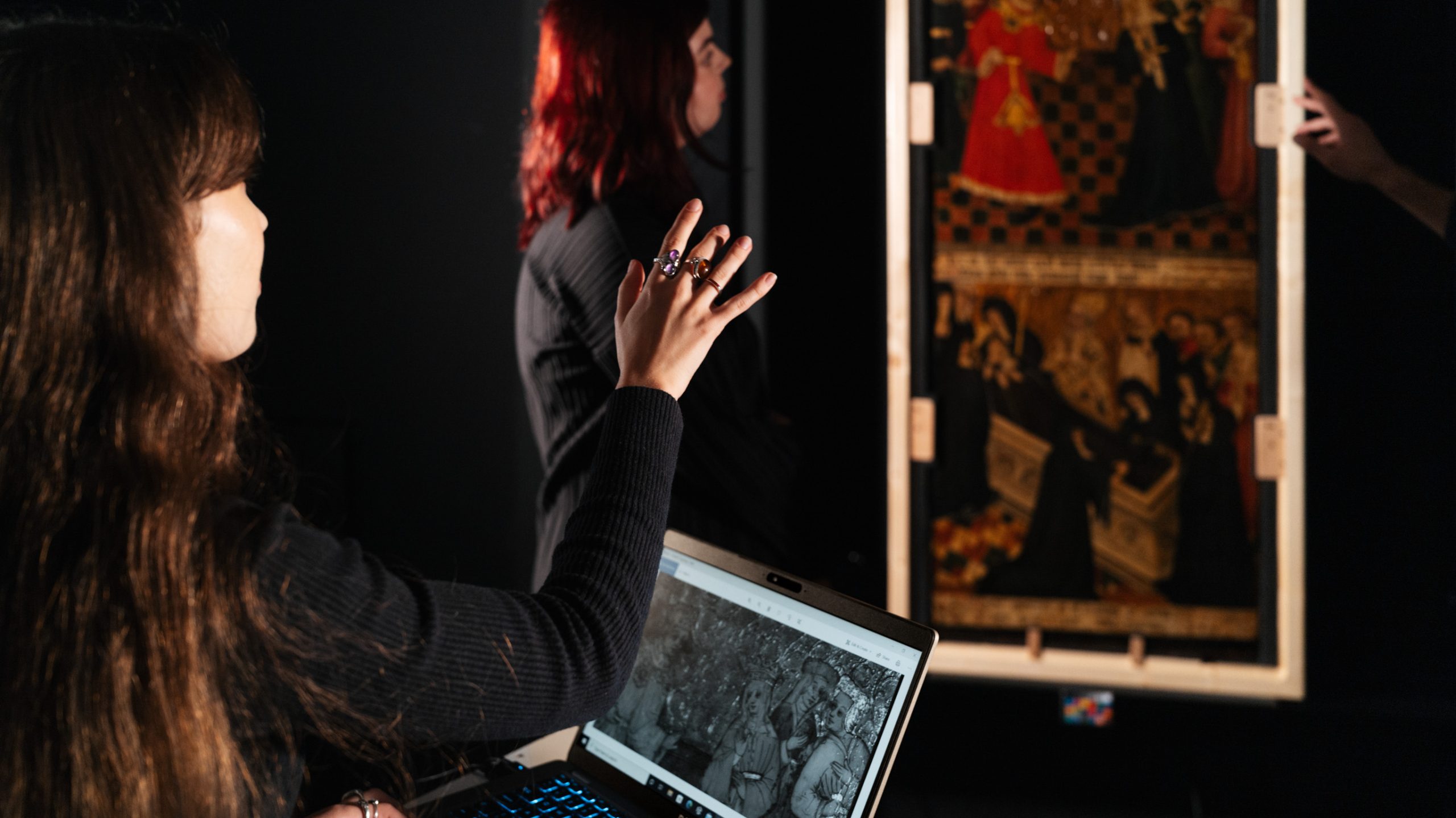 In a dark studio, two women look at an artwork on the wall. One has a laptop open in front of her with a black and white photograph of the same artwork.