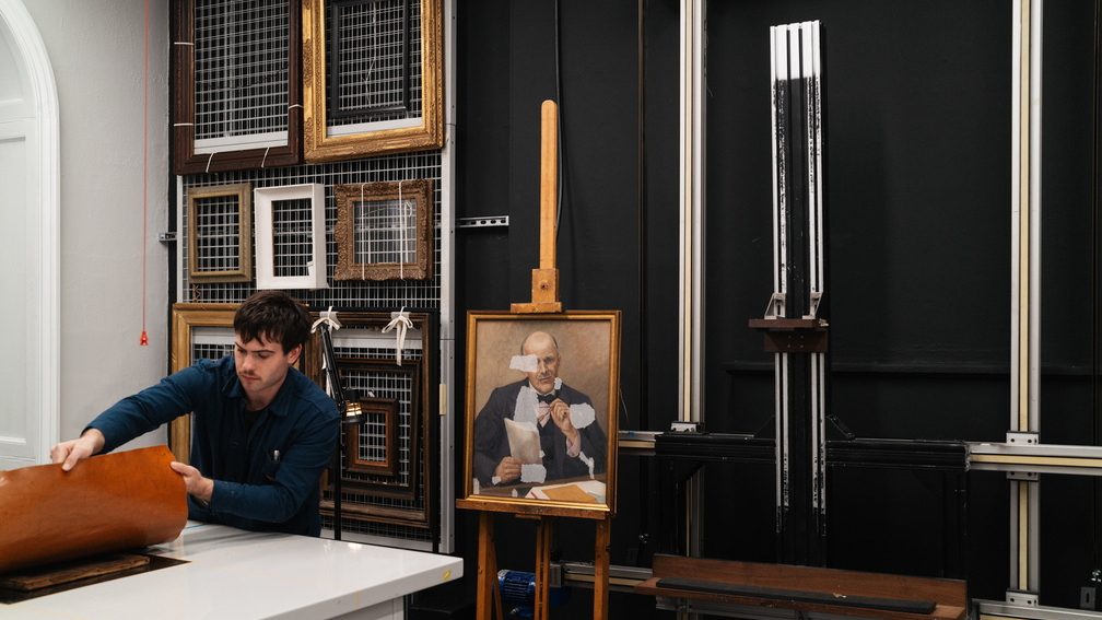 A painting sits on an easel, translucent patches on its surface as if it is being worked on. A man at a table in front of the work lifts a piece of paper off a desk.