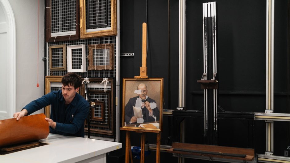 A painting sits on an easel, translucent patches on its surface as if it is being worked on. A man at a table in front of the work lifts a piece of paper off a desk.