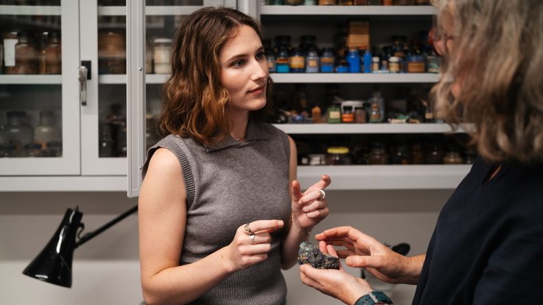 Two women in discussion. One holds a small rock in her palm. Behind them, a shelf of pigments in little antique bottles.
