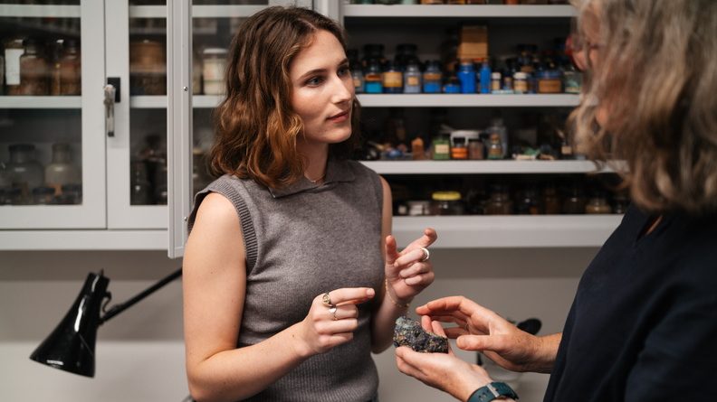 Two women in discussion. One holds a small rock in her palm. Behind them, a shelf of pigments in little antique bottles.