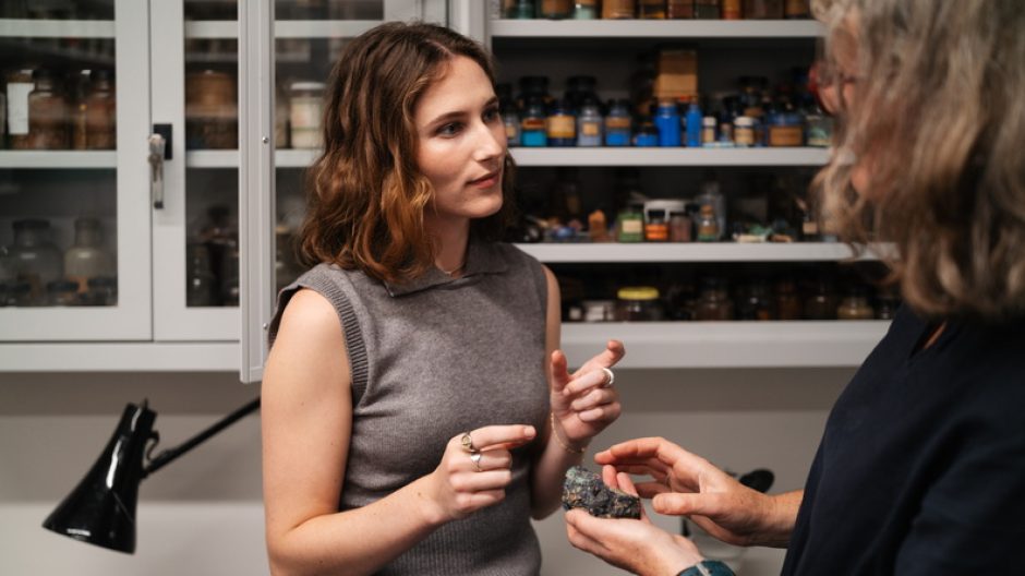 Two women in discussion. One holds a small rock in her palm. Behind them, a shelf of pigments in little antique bottles.
