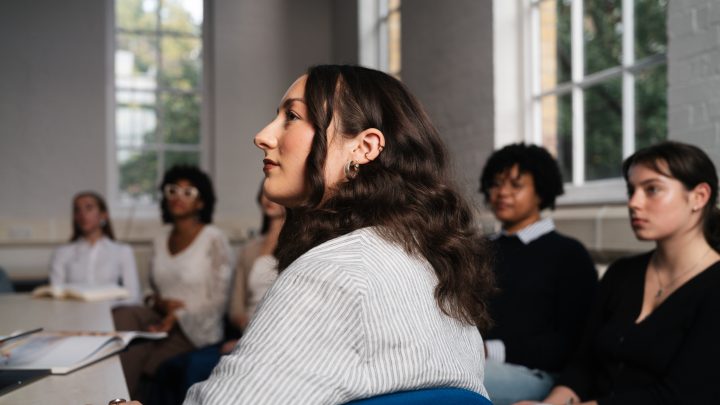 A student sits in a classroom with other students in the background and looks out contemplatively.