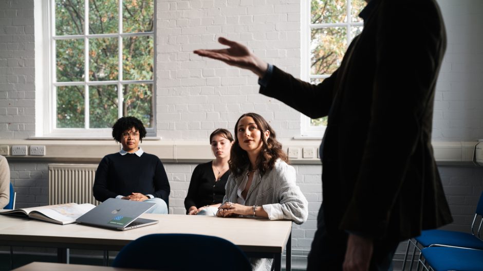 three female students sitting in a seminar room looking up at a tutor who's standing and pointing at something out of sight