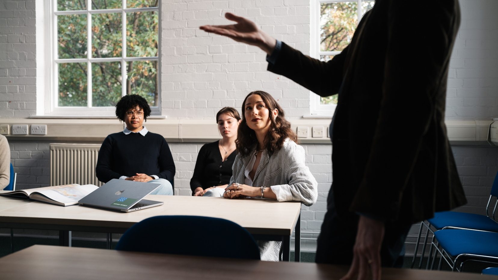 three female students sitting in a seminar room looking up at a tutor who's standing and pointing at something out of sight