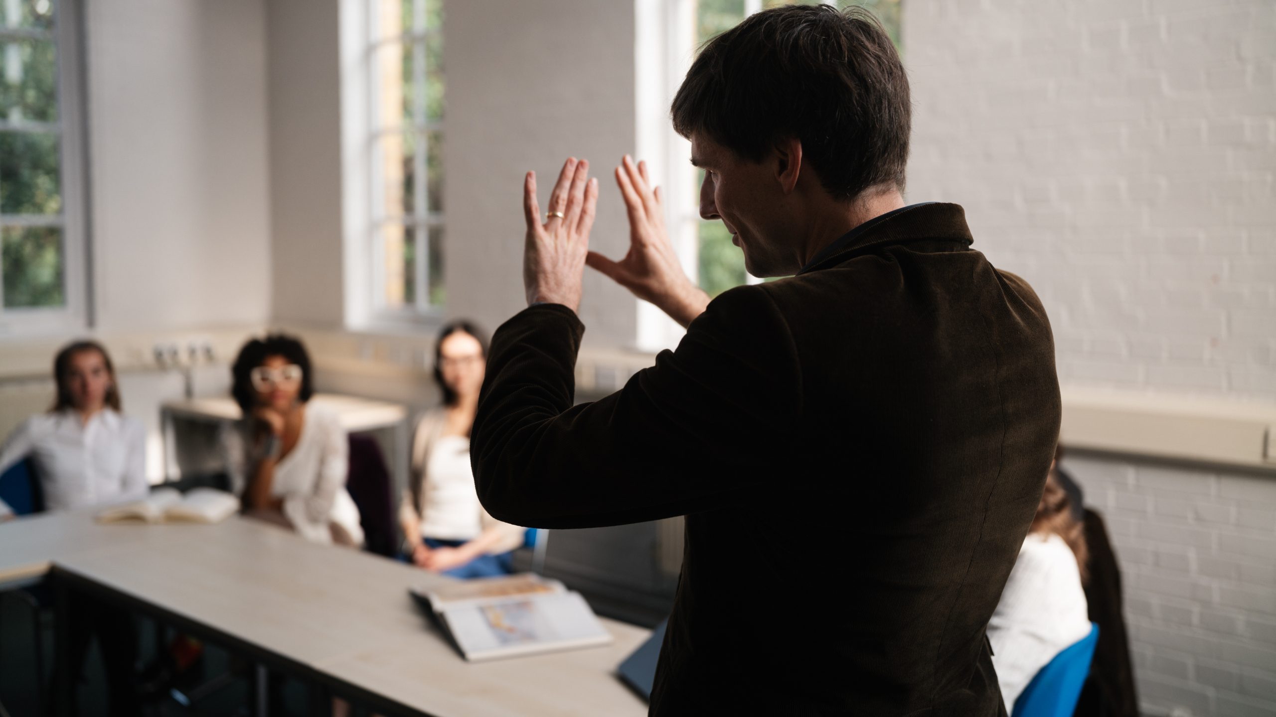 Tutor stands in front of class, his back to the camera. He is gesturing with his hands while students watch on.