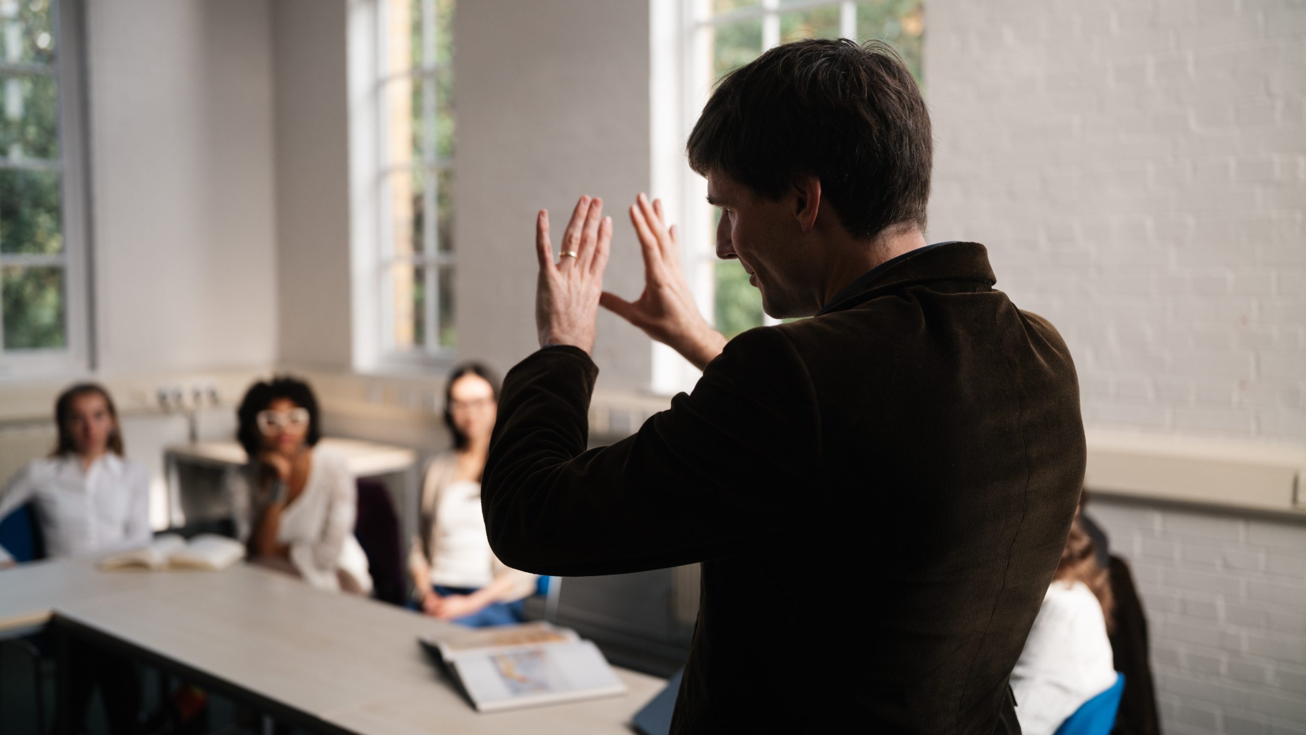 Tutor stands in front of class, his back to the camera. He is gesturing with his hands while students watch on.
