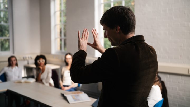 Tutor stands in front of class, his back to the camera. He is gesturing with his hands while students watch on.
