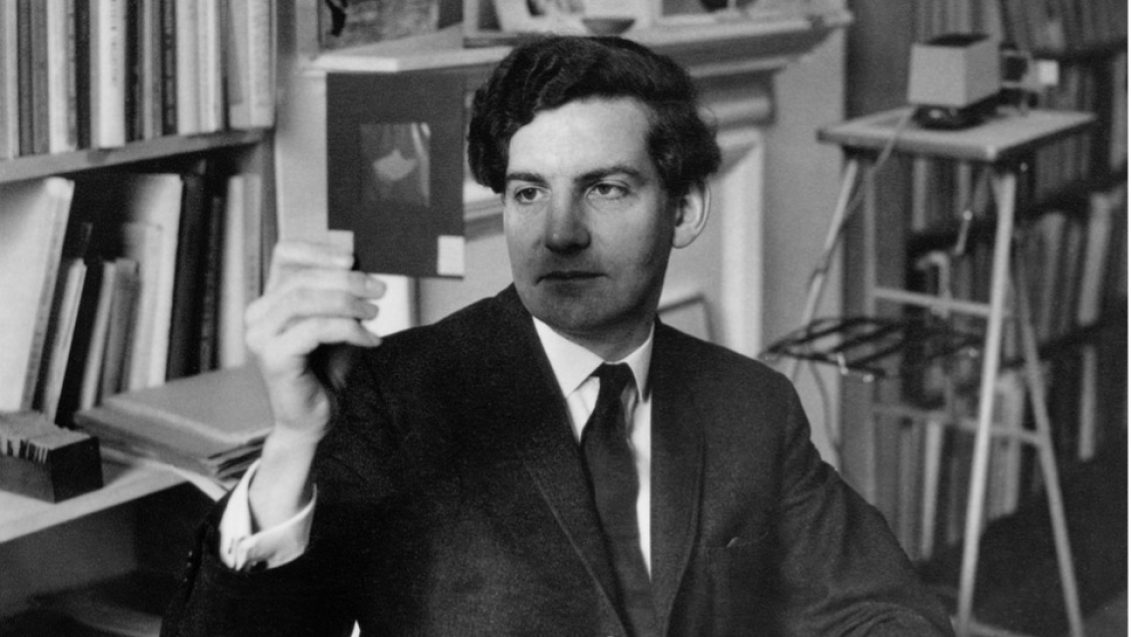 A man in a suit and tie, sat down in an office full of books, looking at an image held up in front of him