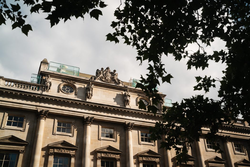 Exterior view of the historic Somerset House stone building with ornate architectural details and statues, partially framed by tree branches.