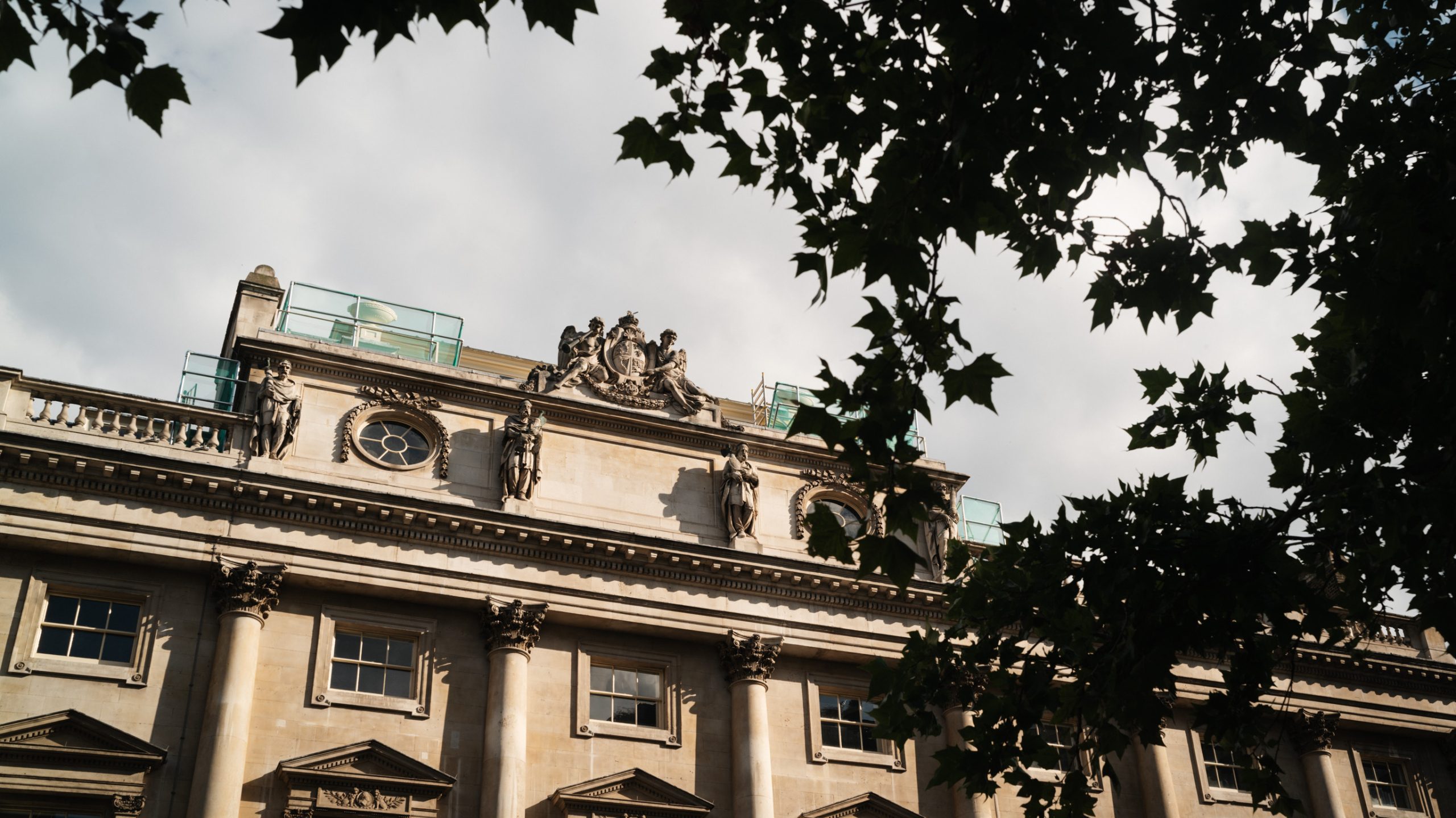 Exterior view of the historic Somerset House stone building with ornate architectural details and statues, partially framed by tree branches.