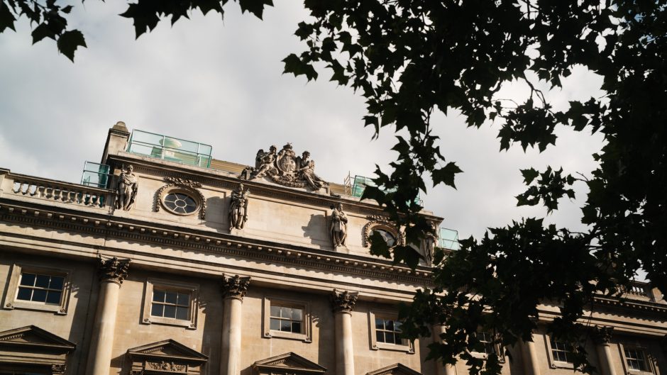 Exterior view of the historic Somerset House stone building with ornate architectural details and statues, partially framed by tree branches.