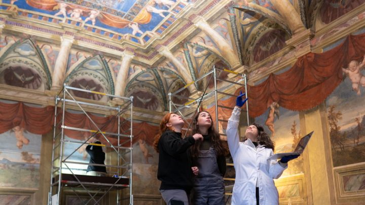 Two students and a teacher, who wears a white lab coat, point at a ceiling in an Italian villa. There is scaffolding up and they are using technical conservation equipment.