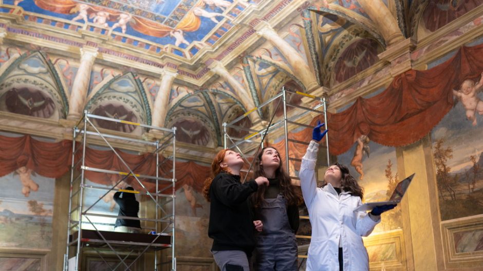 Two students and a teacher, who wears a white lab coat, point at a ceiling in an Italian villa. There is scaffolding up and they are using technical conservation equipment.