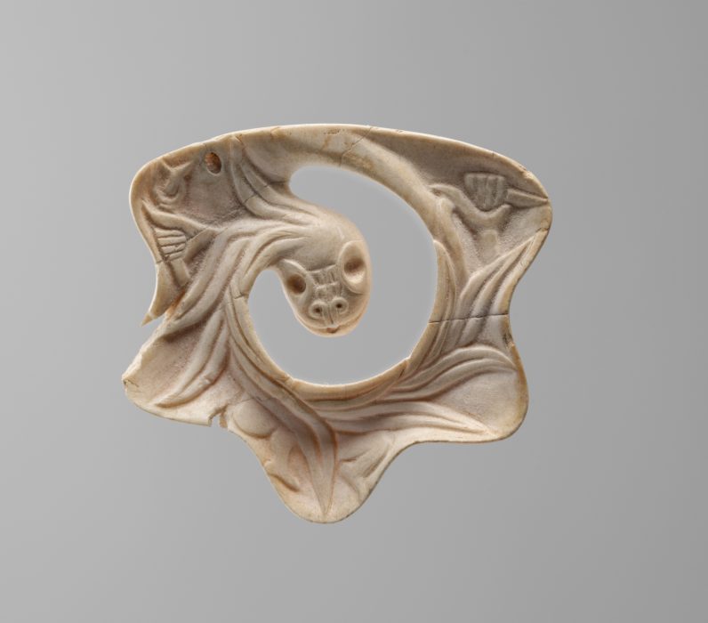 Small Mexica pendant in the shape of the Feathered Serpent, carved from shell with intricate detailing of feathers and serpent features, displaying a polished, slightly curved surface with delicate incisions.