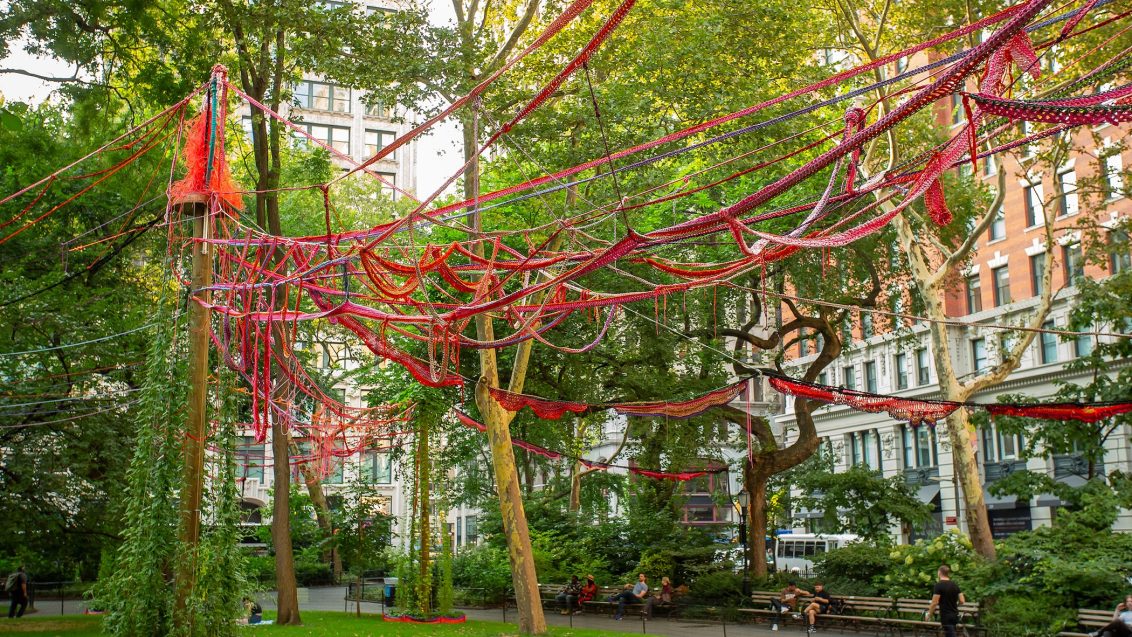Large-scale outdoor installation of interlaced strings, ropes, shoelaces, and plant materials stretched between wooden poles, forming a colorful, web-like structure that evokes a garden canopy within an open park setting.