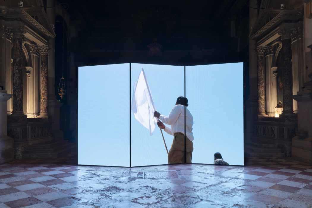 A large three‑panel video screen stands inside an ornate, dimly lit hall. The screen displays a bright image of a person waving a white flag against a clear blue sky. The hall’s marble floor and carved architectural details frame the glowing installation.