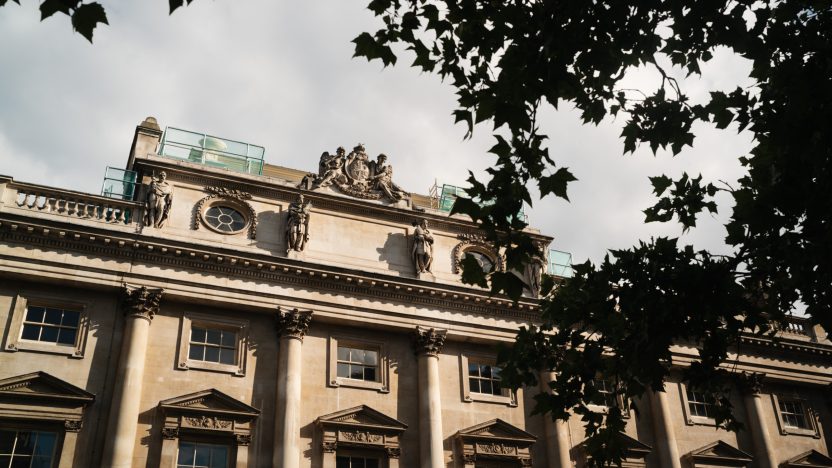 Exterior view of the historic Somerset House stone building with ornate architectural details and statues, partially framed by tree branches.