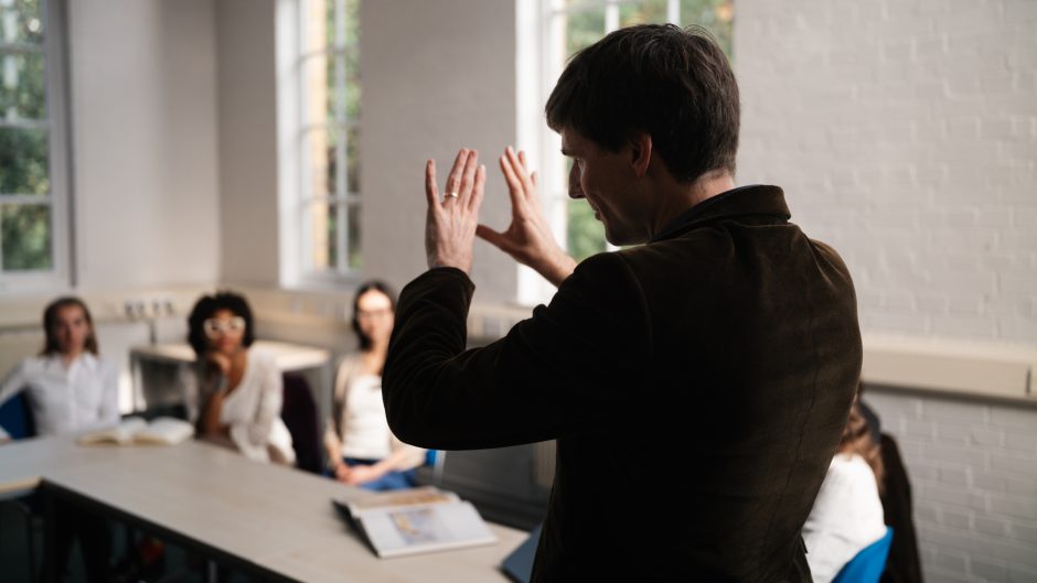 Tutor stands in front of class, his back to the camera. He is gesturing with his hands while students watch on.