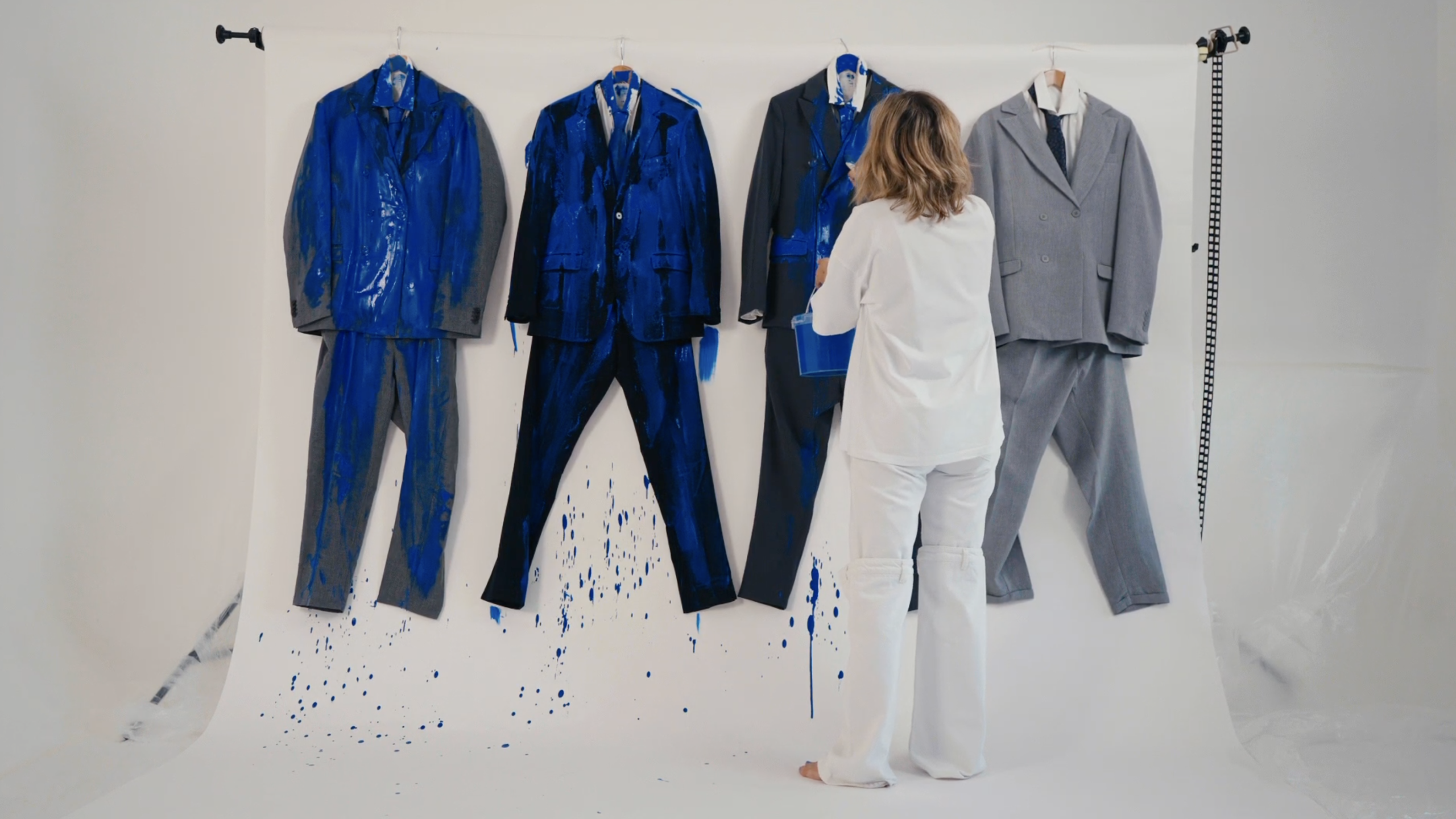 A person wearing white clothes stands in a photo studio, painting men’s suits that hang on a white backdrop. Four suits are arranged in a row on hangers; three of them are splattered with bright blue paint, which drips onto the backdrop and floor. The person is seen from behind, holding a paint bucket and brush as they apply more blue paint to one of the suits. The space is brightly lit and minimalist.