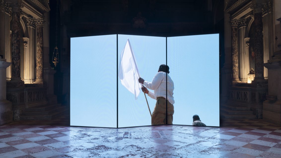 A large three‑panel video screen stands inside an ornate, dimly lit hall. The screen displays a bright image of a person waving a white flag against a clear blue sky. The hall’s marble floor and carved architectural details frame the glowing installation.