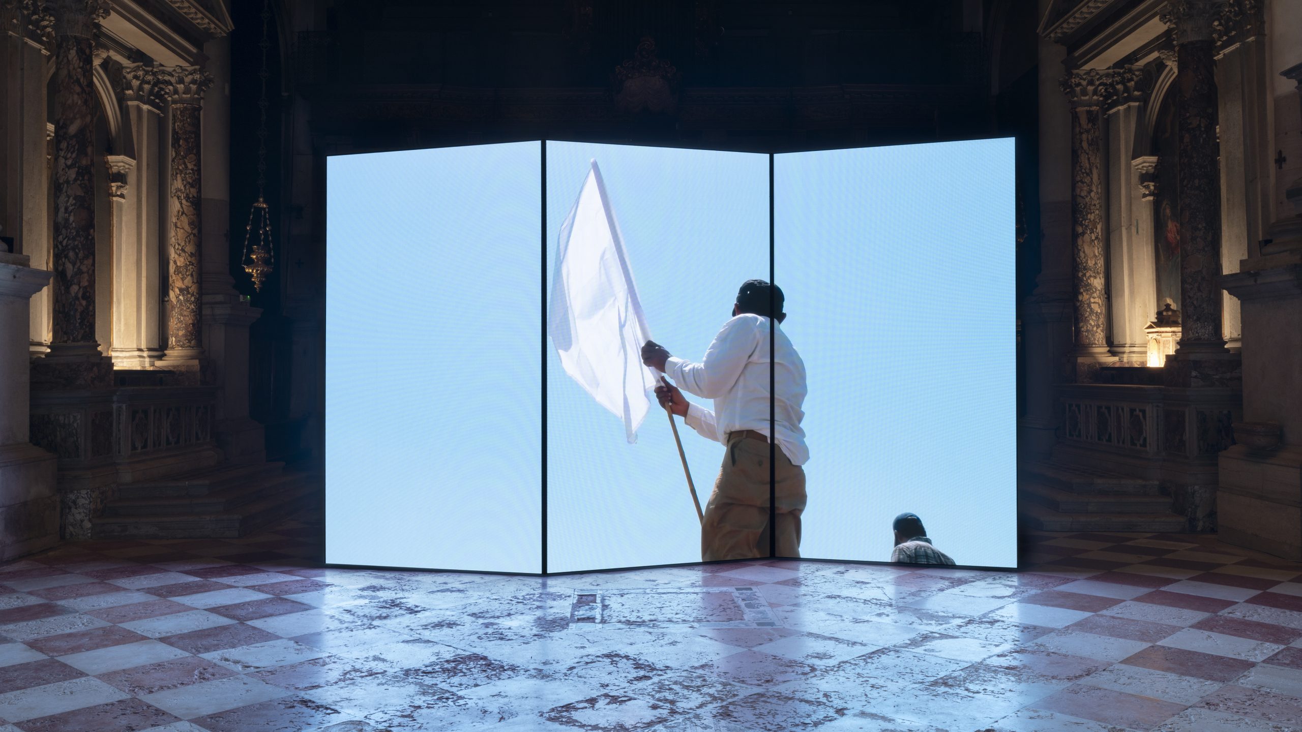 A large three‑panel video screen stands inside an ornate, dimly lit hall. The screen displays a bright image of a person waving a white flag against a clear blue sky. The hall’s marble floor and carved architectural details frame the glowing installation.