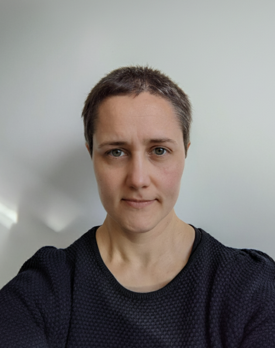 A white woman with short hair looking straight on to the camera against a grey background. She wears a dark navy jumper.