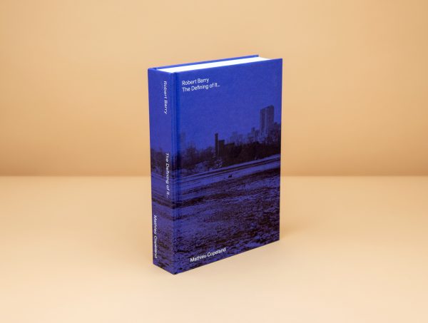 Book cover of Robert Barry, The Defining of It... depicting a snowed empty park in the foreground and a city in the background. The whole photograph has a blue tint over it.