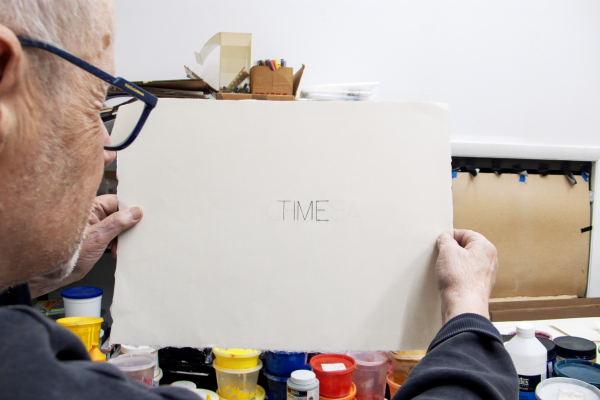 Photograph of Robert Barry in his studio, holding a piece of white paper to the camera with the word TIME on it.