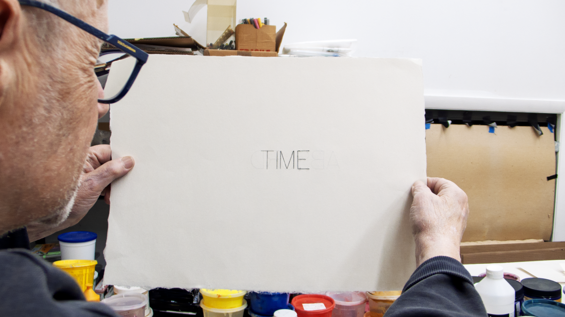 Photograph of Robert Barry in his studio, holding a piece of white paper to the camera with the word TIME on it.