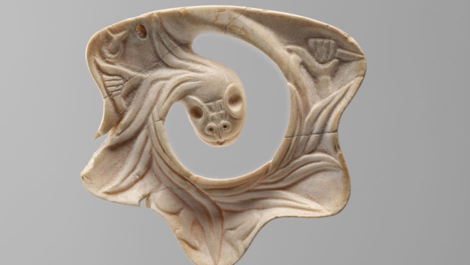 Small Mexica pendant in the shape of the Feathered Serpent, carved from shell with intricate detailing of feathers and serpent features, displaying a polished, slightly curved surface with delicate incisions.