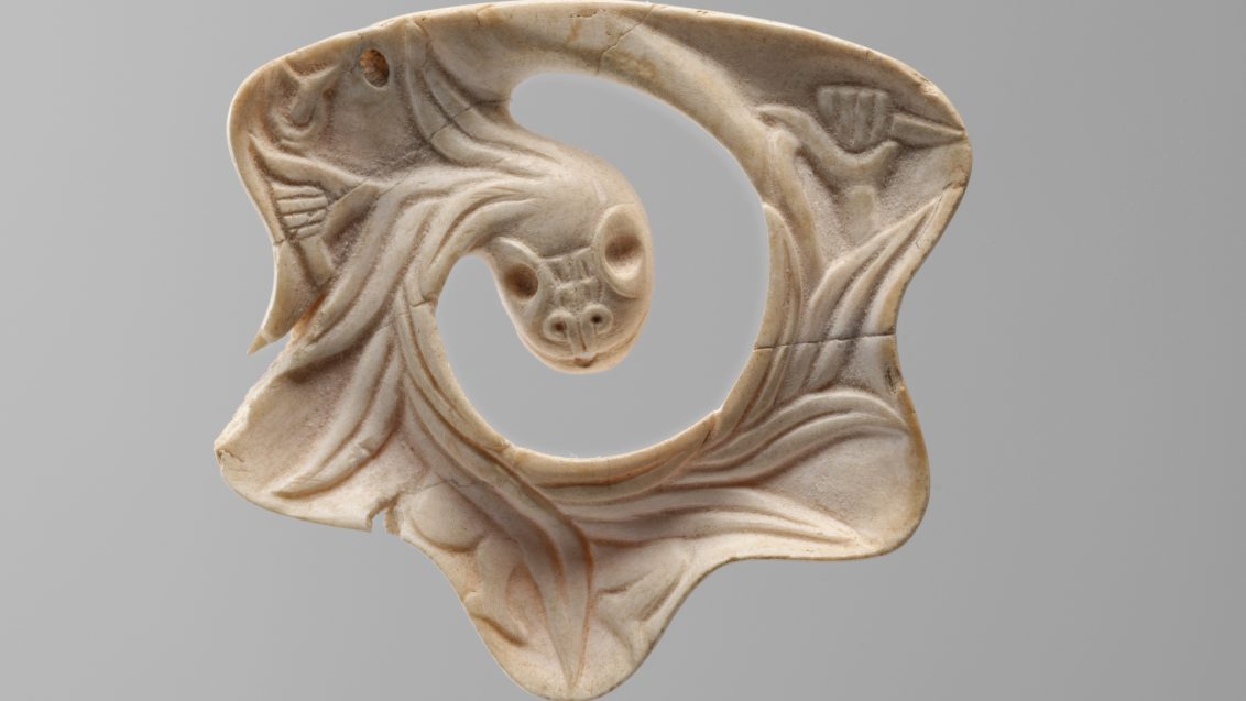 Small Mexica pendant in the shape of the Feathered Serpent, carved from shell with intricate detailing of feathers and serpent features, displaying a polished, slightly curved surface with delicate incisions.