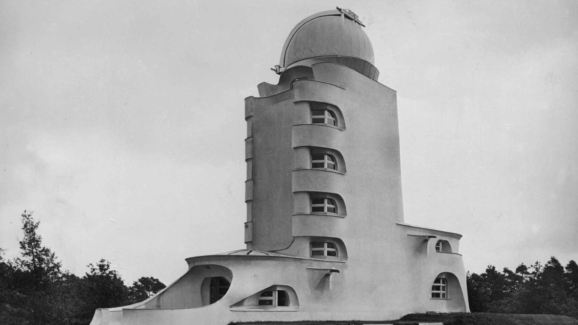 Black‑and‑white photograph of a tall, curved observatory building with smooth, sculptural walls and a domed roof. The structure features narrow, rounded windows arranged in a vertical line and sits in a grassy landscape bordered by trees.