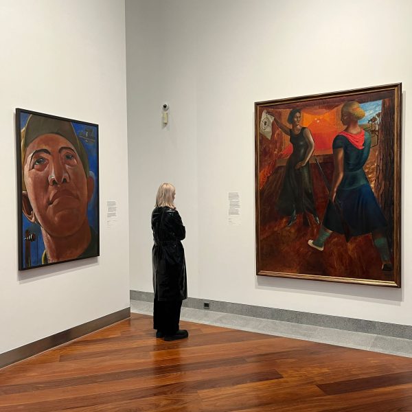 A woman stands in a gallery where two walls meet; she is between two paintings looking at one