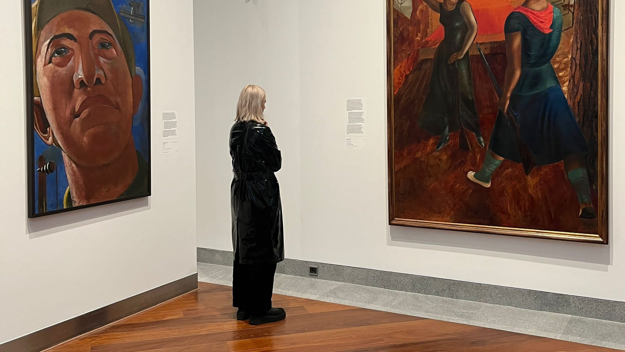 A woman stands in a gallery where two walls meet; she is between two paintings looking at one