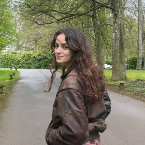 A woman with long brown hair wearing a brown leather jacket, walking in a park, turns to look at the camera and smiles