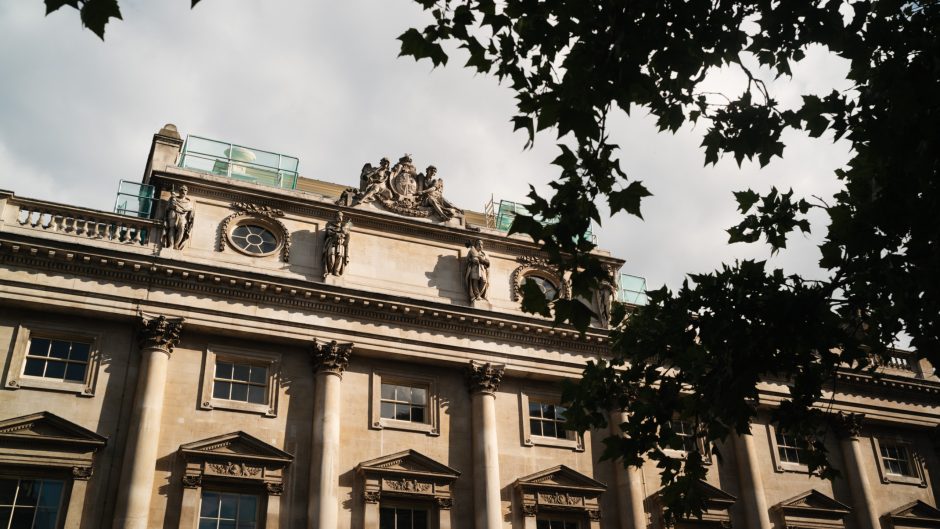 Exterior view of the historic Somerset House stone building with ornate architectural details and statues, partially framed by tree branches.