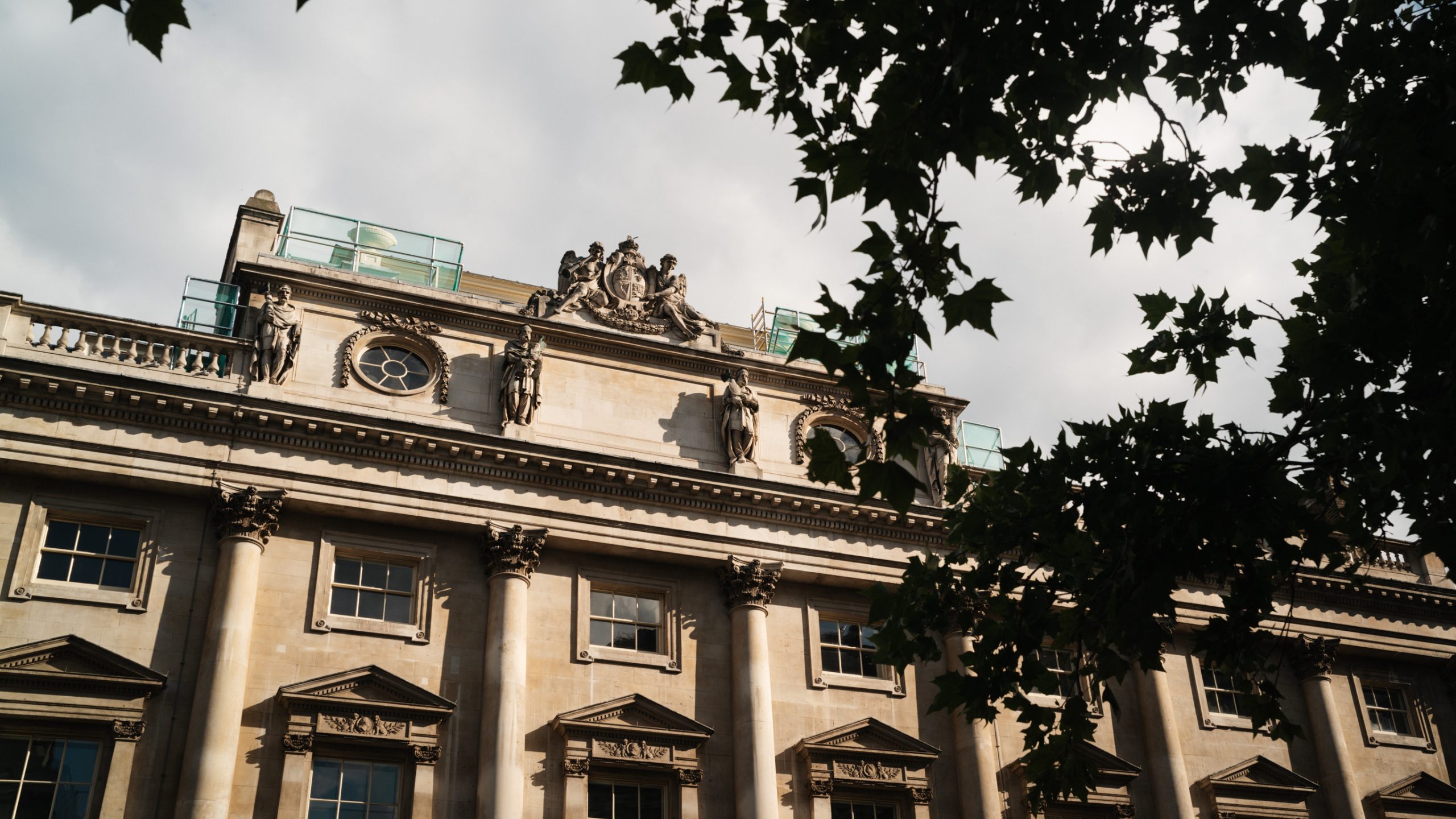 Exterior view of the historic Somerset House stone building with ornate architectural details and statues, partially framed by tree branches.