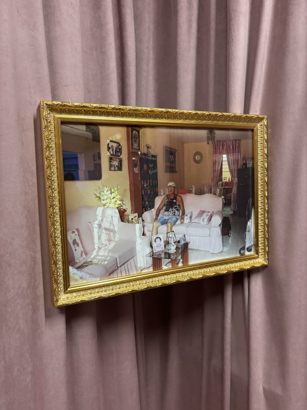 A photograph in a frame hangs on a curtained wall. The photograph depicts a pink sitting room with family photographs and other familial clutter. An elderly man in a cap sits on a sofa gazing out of frame.